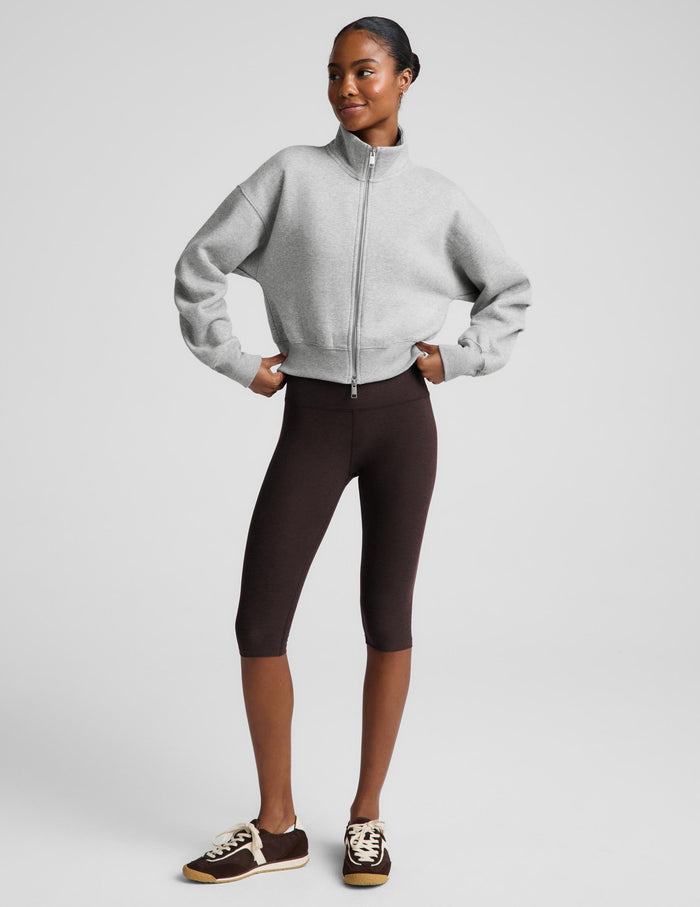 A woman modeling a light gray zip-up fleece jacket and dark brown capri leggings against a light gray studio background.