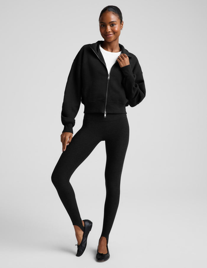 A woman wearing a black cropped zip-up sweatshirt with a stand collar over a white top and black leggings, photographed against a plain light gray studio background.