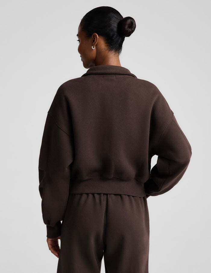 A woman wearing a dark brown zip-up sweatshirt and matching sweatpants against a light gray studio background.