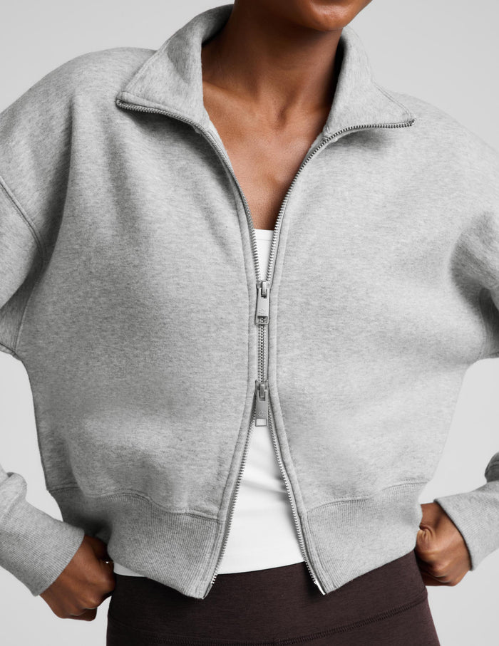A woman modeling a light gray zip-up fleece jacket and dark brown capri leggings against a light gray studio background.