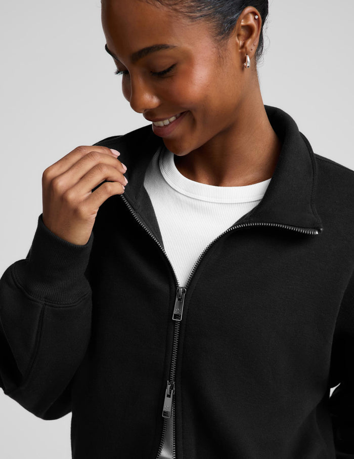 A woman wearing a black cropped zip-up sweatshirt with a stand collar over a white top and black leggings, photographed against a plain light gray studio background.