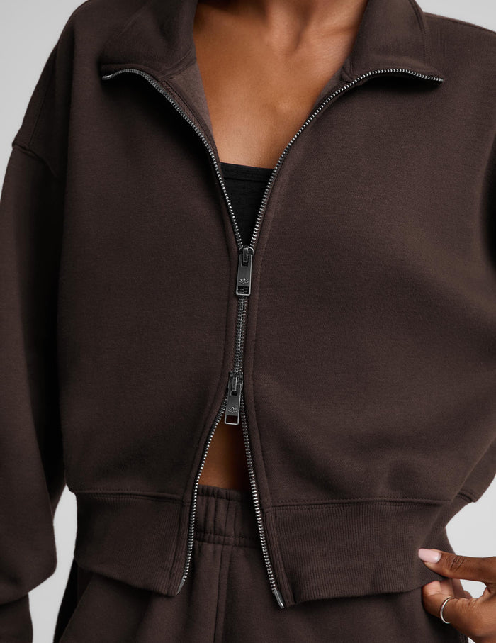 A woman wearing a dark brown zip-up sweatshirt and matching sweatpants against a light gray studio background.