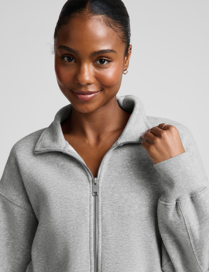 A woman modeling a light gray zip-up fleece jacket and dark brown capri leggings against a light gray studio background.