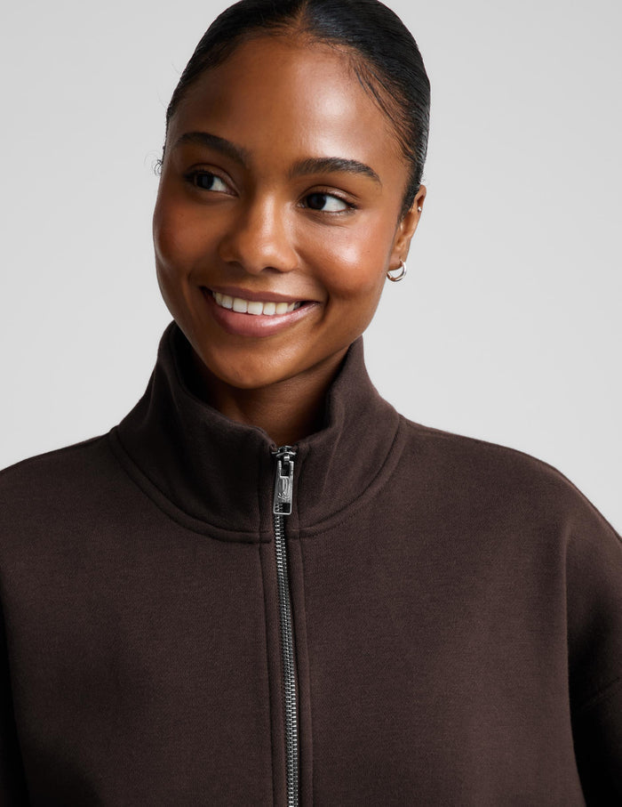 A woman wearing a dark brown zip-up sweatshirt and matching sweatpants against a light gray studio background.