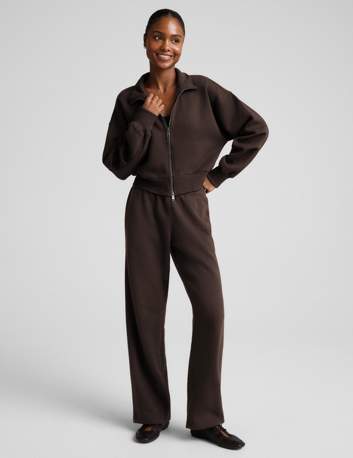 A woman wearing a dark brown zip-up sweatshirt and matching sweatpants against a light gray studio background.