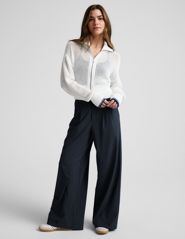 Woman wearing a white zip-up sweater and blue trousers against a plain background.