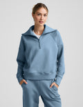 Woman wearing a light blue quarter zip pullover and pants against a plain background.