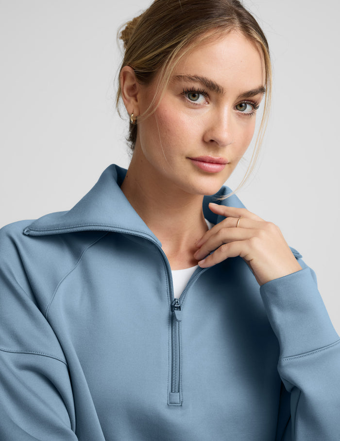 Woman wearing a light blue quarter zip pullover and pants against a plain background.