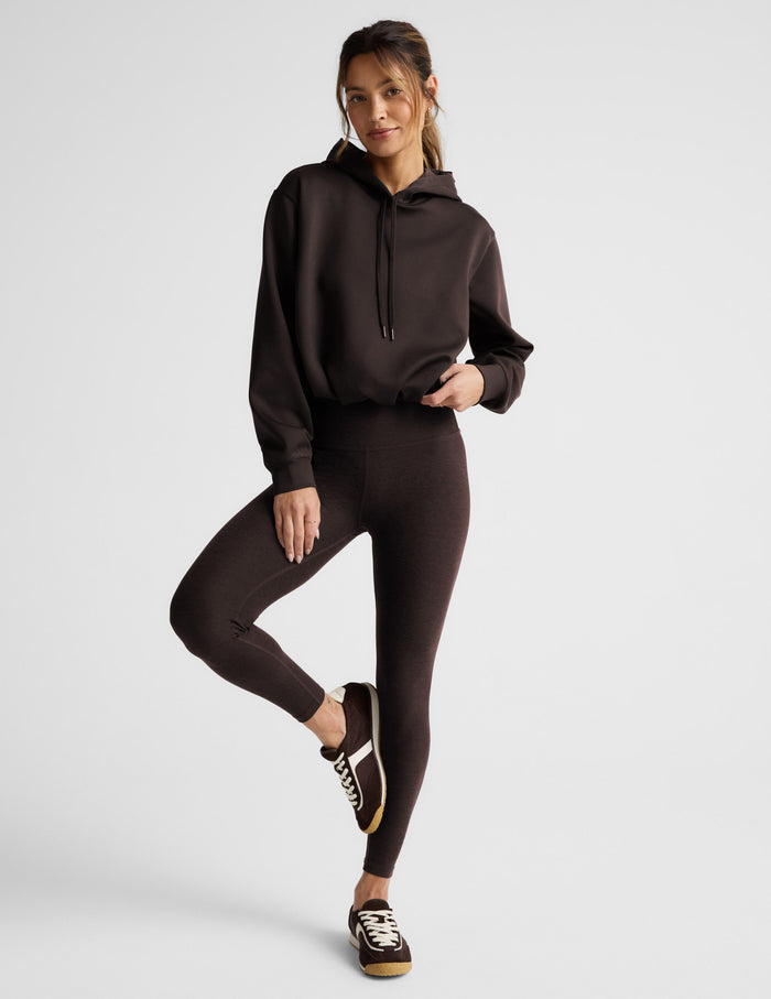 Woman wearing a brown hoodie and leggings on a light gray background.