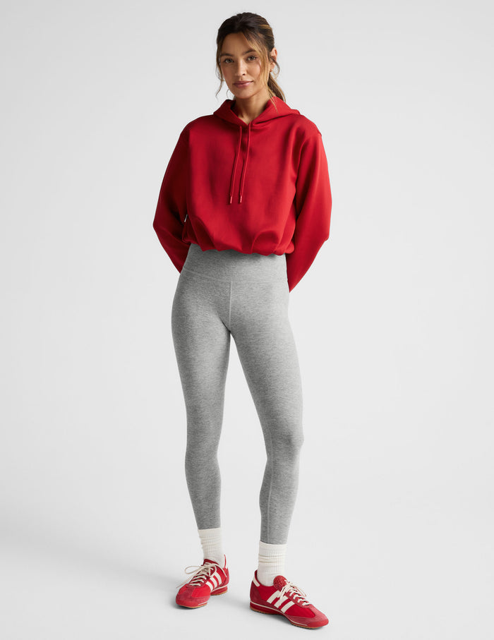 Woman wearing a red hoodie and gray leggings on a light gray background.