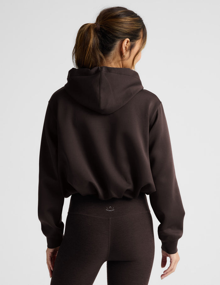 Woman wearing a brown hoodie and leggings on a light gray background.
