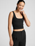 Woman is wearing a black ribbed tank and pants against a light gray background.