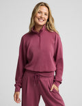 A woman modeling a matching dusty rose half-zip sweatshirt and drawstring sweatpants set against a solid light gray background.