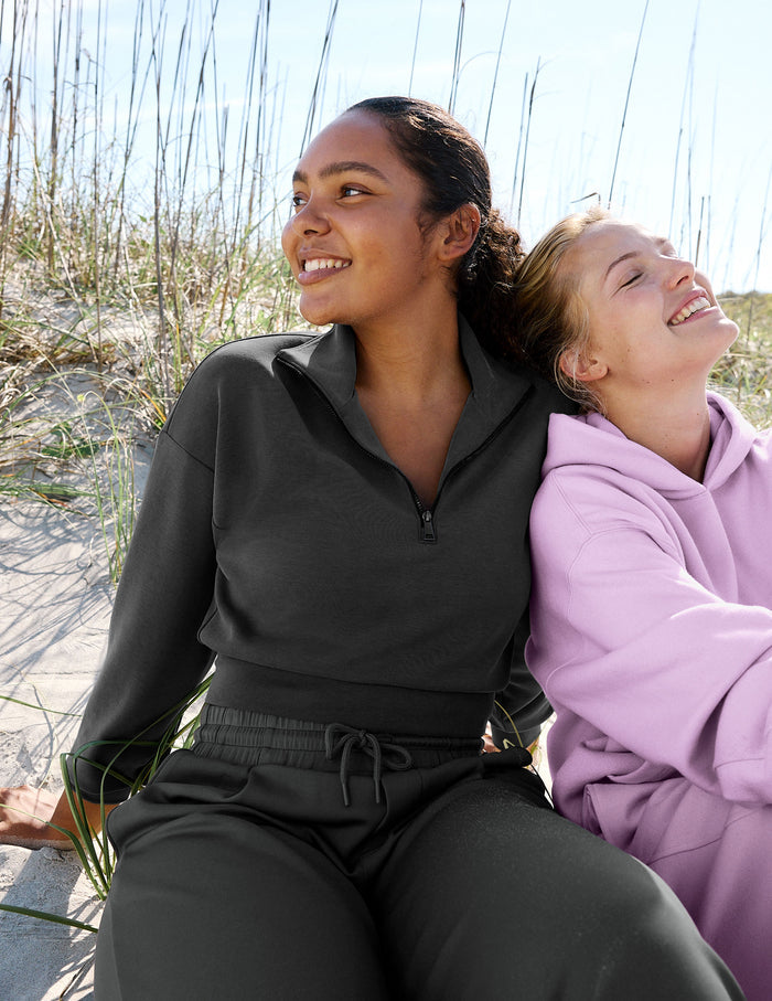 Two women sitting together outdoors on sand dunes, one wearing a dark charcoal gray quarter-zip pullover and matching pants, and the other wearing a light lavender hoodie.