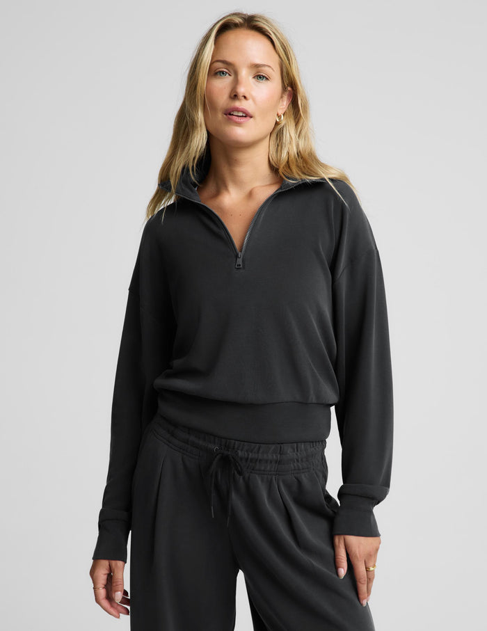 A woman modeling a black long-sleeve quarter-zip pullover top with black sweatpants against a light gray studio background.
