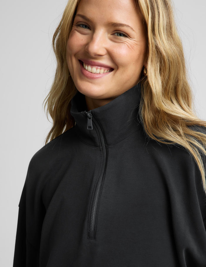 A close-up of a smiling blonde woman wearing a black quarter-zip pullover with a high mock neck collar, set against a plain light gray studio background.