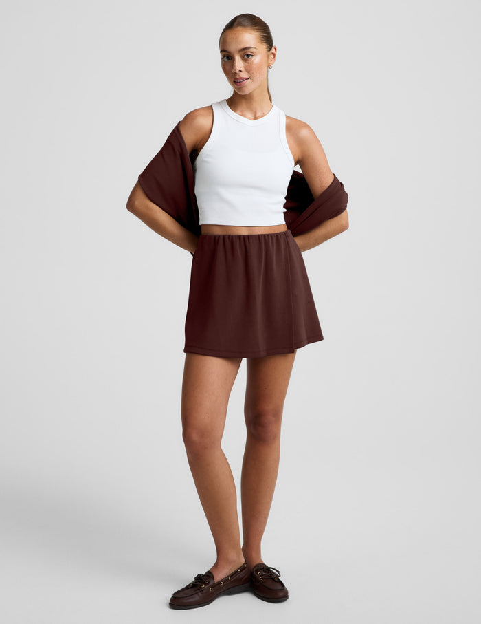 Woman wearing brown Luxe Sandwash™ Skort with a white ribbed tank top and a brown sweater draped over her shoulders.