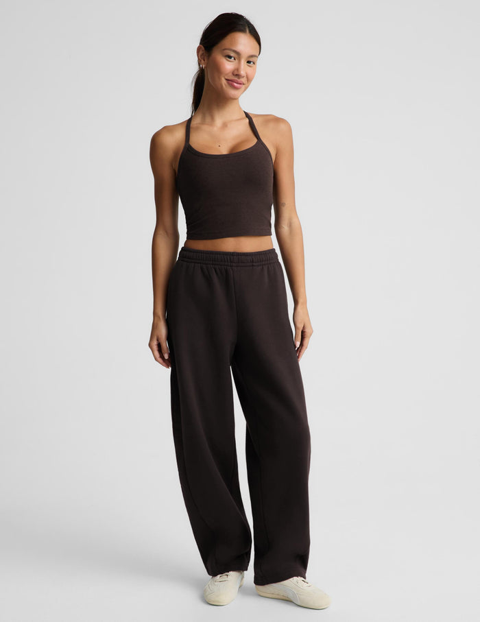 A woman modeling a dark brown activewear set consisting of a crop tank and matching barrel leg sweatpants, paired with white sneakers, against a light gray studio background.