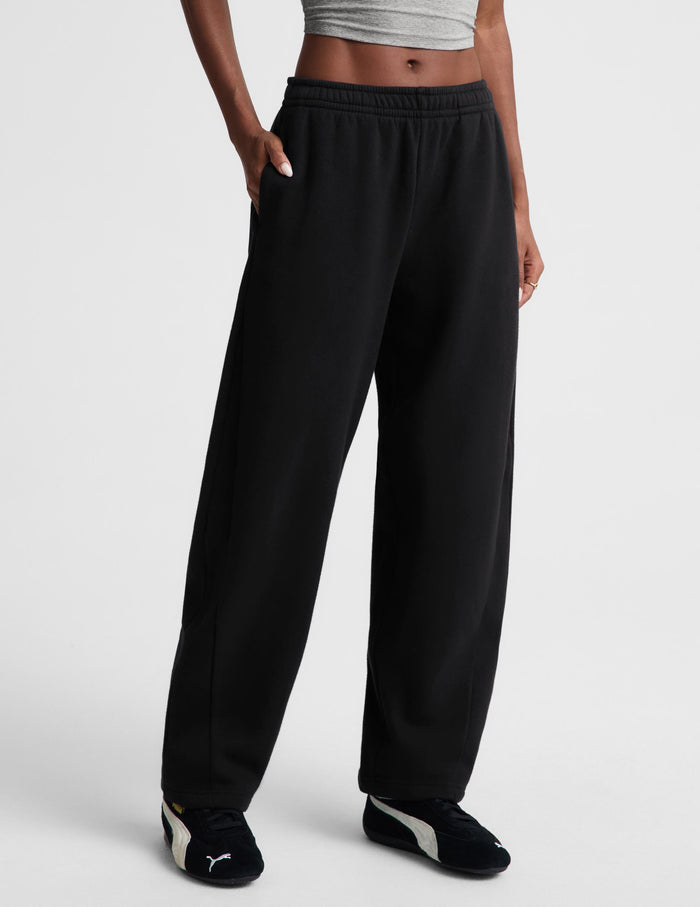 A person wearing black wide-leg sweatpants with an elastic waistband, paired with a gray crop top and black-and-white Puma sneakers, shown against a light gray studio background.