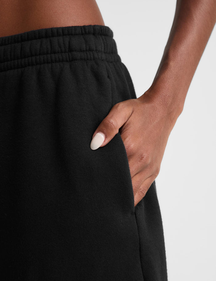 Close-up of a person wearing black sweatpants, showing the side pocket and hand with white manicured nails resting in the pocket against a light gray background.