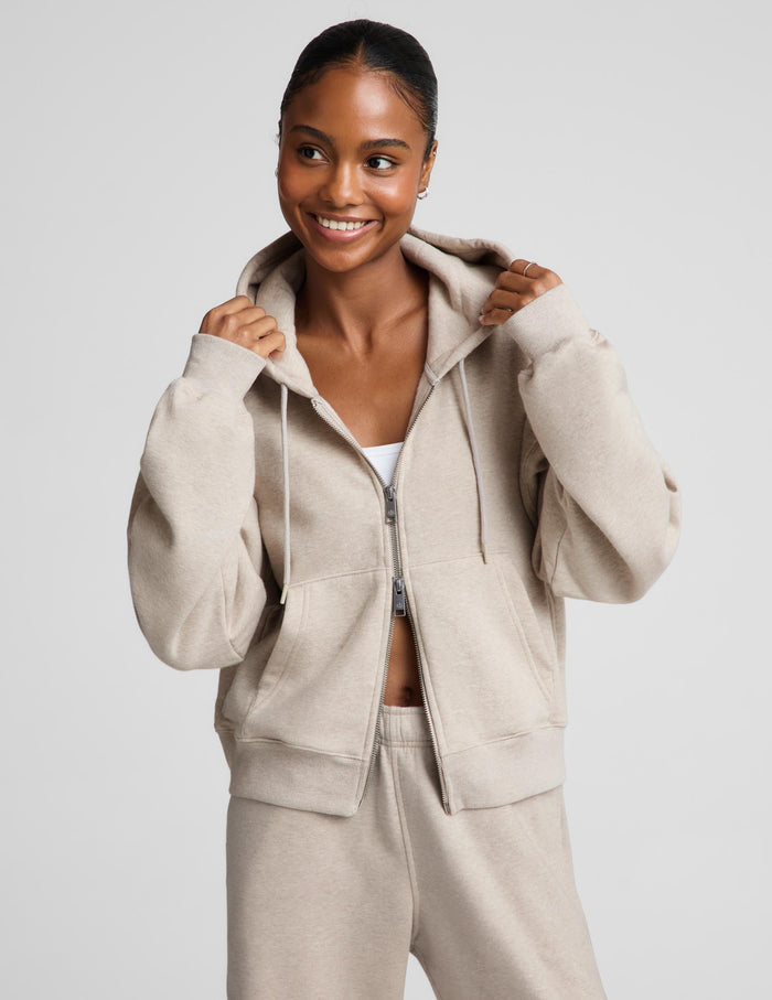 A woman wearing a light beige zip-up hoodie and matching sweatpants, posing against a solid light gray background.