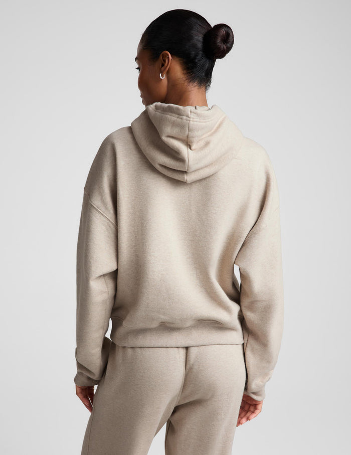 A woman shown from the back wearing a light beige hooded sweatshirt and matching sweatpants against a plain light gray studio background.