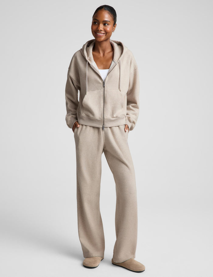 A woman shown from the back wearing a light beige hooded sweatshirt and matching sweatpants against a plain light gray studio background.