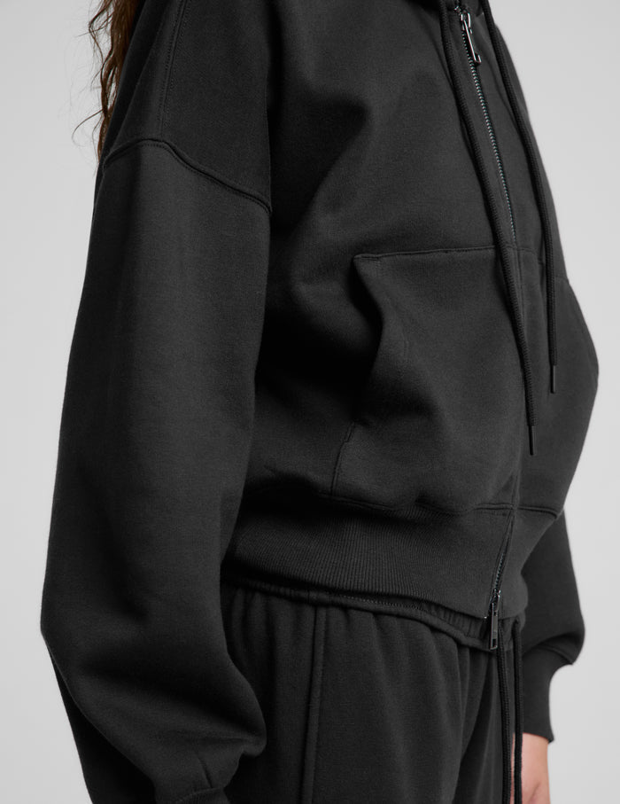 Woman wearing a black fleece zip-up sweatshirt and black sweatpants against a plain background.