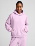 A woman wearing a light pink matching lounge set, featuring a pullover hoodie with a front kangaroo pocket and matching sweatpants, set against a solid light gray background.