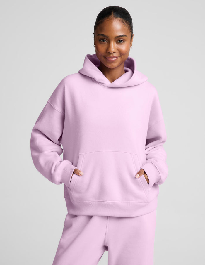 A woman wearing a light pink matching lounge set, featuring a pullover hoodie with a front kangaroo pocket and matching sweatpants, set against a solid light gray background.