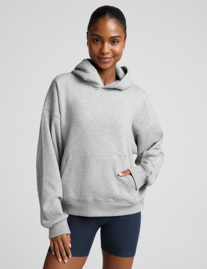 A woman wearing an oversized light gray heather pullover hoodie with a front kangaroo pocket, paired with navy blue bike shorts, against a solid light gray studio background.