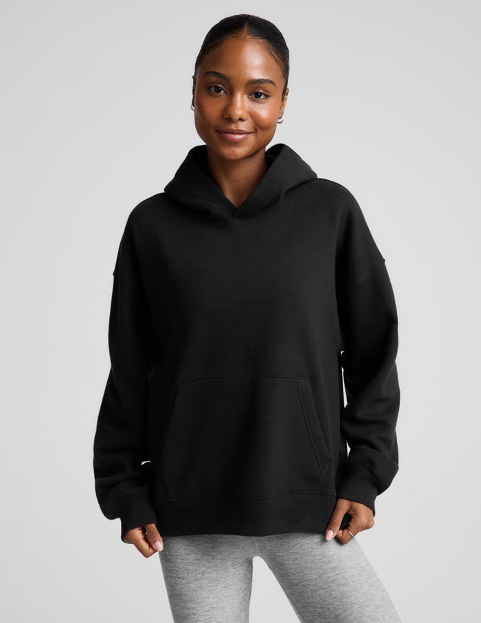 A woman modeling a black pullover hoodie with a front kangaroo pocket, paired with light gray sweatpants, against a solid light gray background.