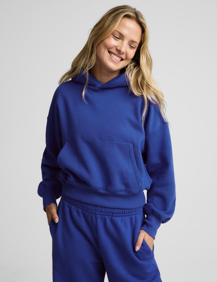 A woman wearing a matching royal blue sweatshirt and jogger pants set, smiling with her eyes closed against a light gray studio background.