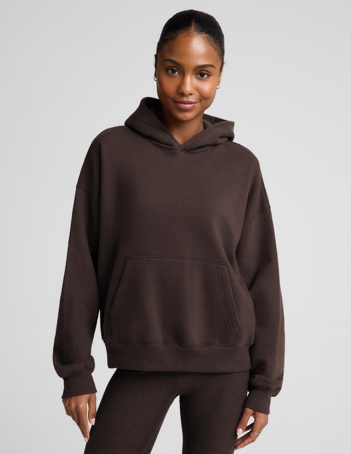 A woman wearing a dark brown pullover hoodie with a front kangaroo pocket, paired with matching dark brown leggings, against a solid light gray studio background.