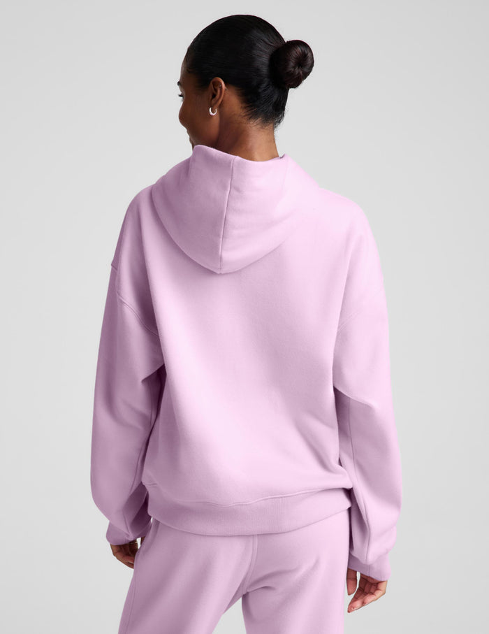 A woman wearing a light pink matching lounge set, featuring a pullover hoodie with a front kangaroo pocket and matching sweatpants, set against a solid light gray background.
