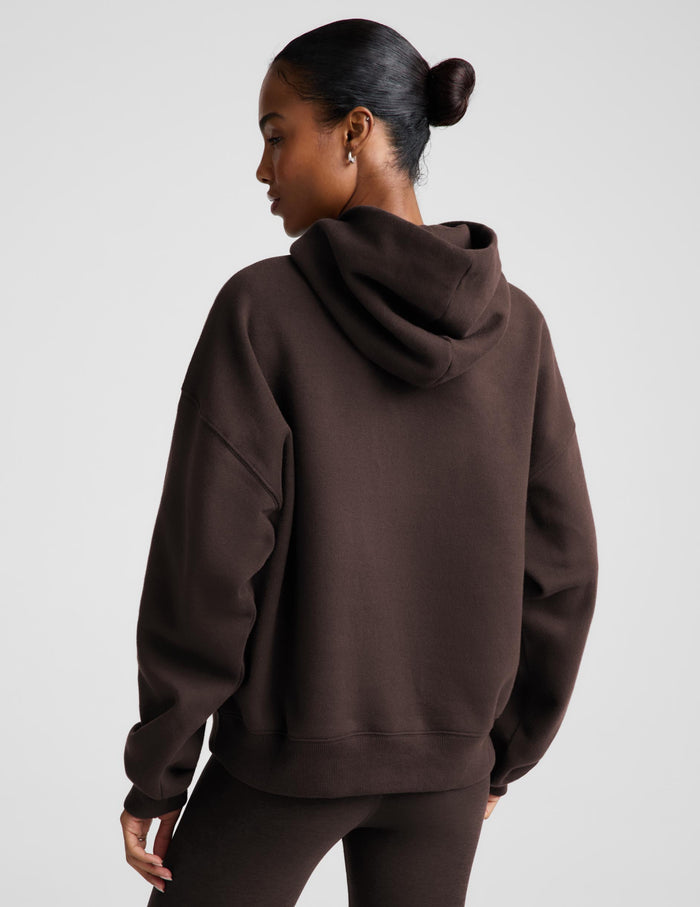 A woman wearing a dark brown pullover hoodie with a front kangaroo pocket, paired with matching dark brown leggings, against a solid light gray studio background.