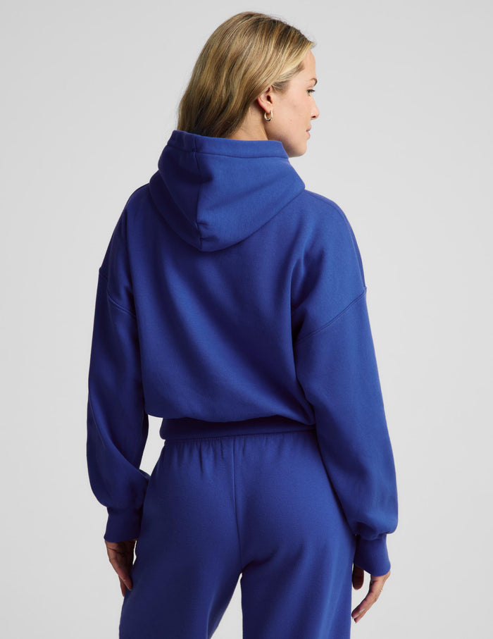 A woman shown from the back wearing a royal blue cropped hoodie and matching sweatpants against a light gray studio background.
