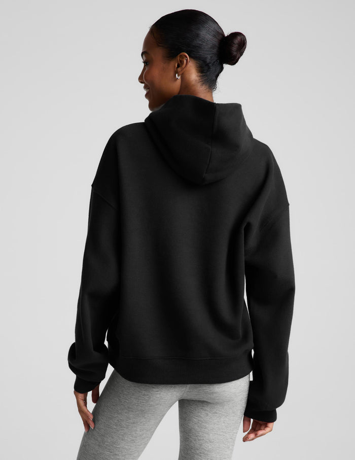 A woman modeling a black pullover hoodie with a front kangaroo pocket, paired with light gray leggings, against a solid light gray background.