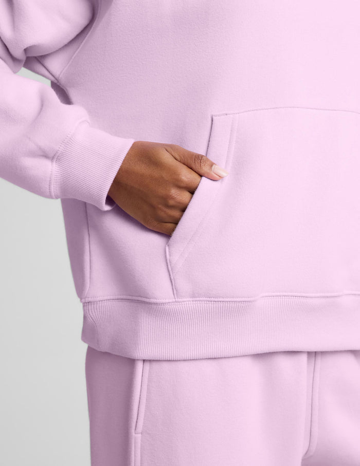 A woman wearing a light pink matching lounge set, featuring a pullover hoodie with a front kangaroo pocket and matching sweatpants, set against a solid light gray background.