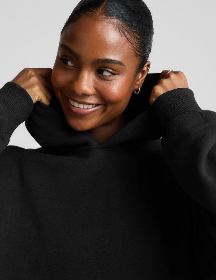 A woman modeling a black pullover hoodie with a front kangaroo pocket, paired with light gray leggings, against a solid light gray background.