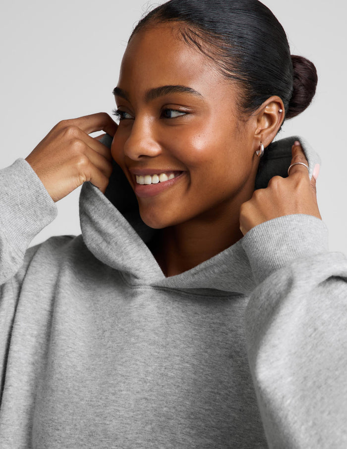 A woman wearing an oversized light gray heather pullover hoodie with a front kangaroo pocket, paired with navy blue bike shorts, against a solid light gray studio background.