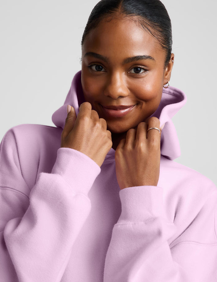 A woman wearing a light pink matching lounge set, featuring a pullover hoodie with a front kangaroo pocket and matching sweatpants, set against a solid light gray background.