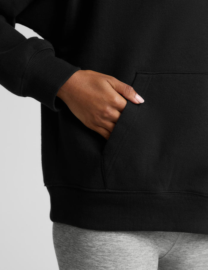 A woman modeling a black pullover hoodie with a front kangaroo pocket, paired with light gray leggings, against a solid light gray background.