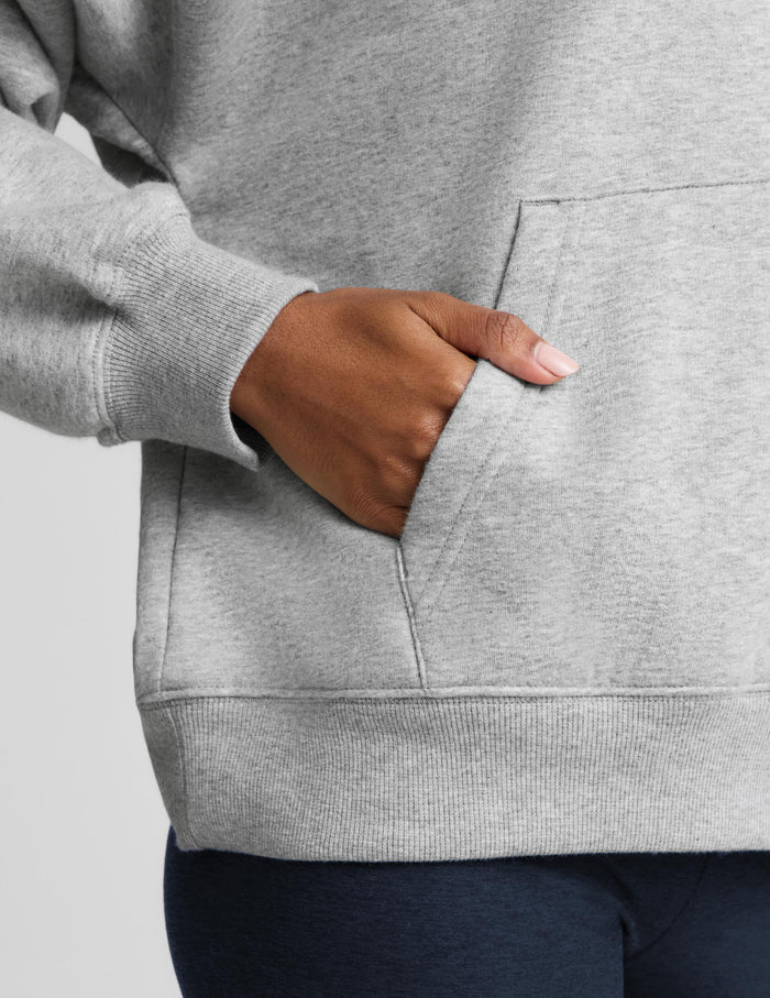 A woman wearing an oversized light gray heather pullover hoodie with a front kangaroo pocket, paired with navy blue bike shorts, against a solid light gray studio background.
