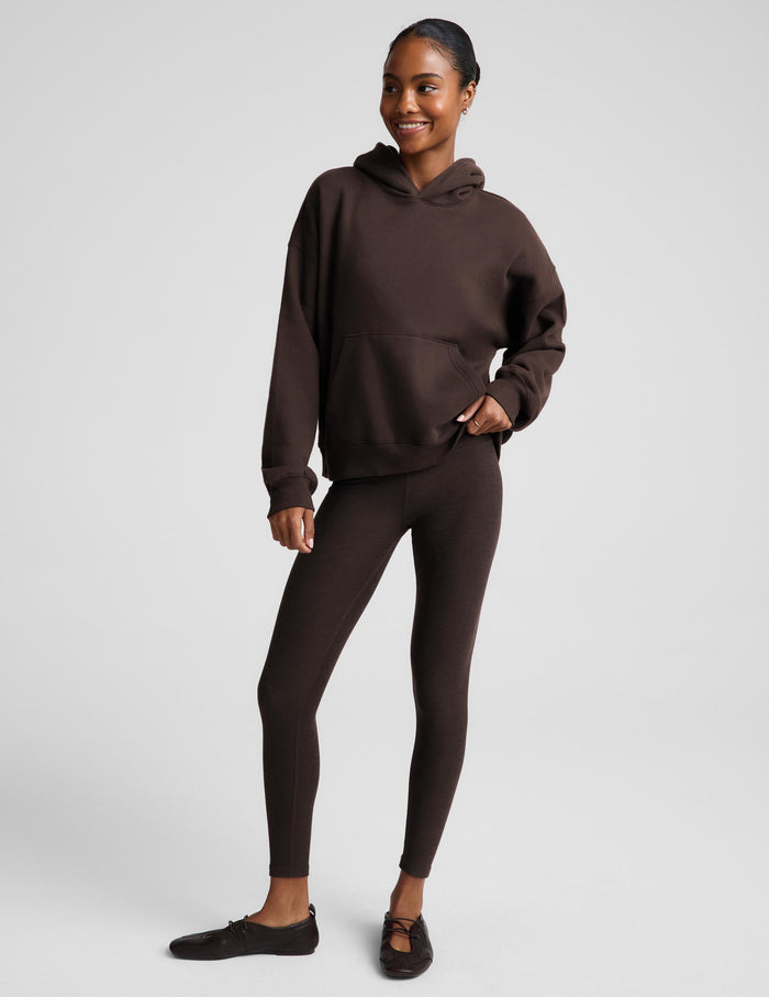 A woman wearing a dark brown pullover hoodie with a front kangaroo pocket, paired with matching dark brown leggings, against a solid light gray studio background.