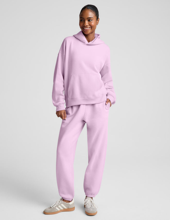 A woman wearing a light pink matching lounge set, featuring a pullover hoodie with a front kangaroo pocket and matching sweatpants, set against a solid light gray background.