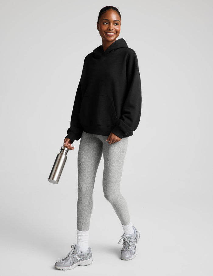 A woman modeling a black pullover hoodie with a front kangaroo pocket, paired with light gray leggings, against a solid light gray background.