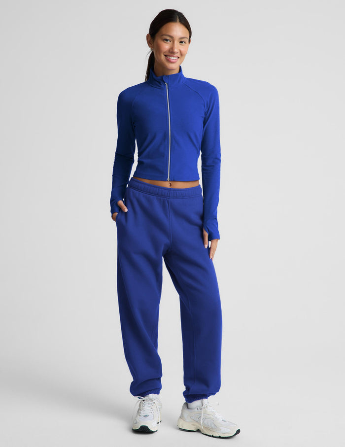 A woman modeling a matching royal blue athletic set consisting of a long-sleeve zip-up and high-waisted sweatpants, paired with white sneakers, against a light gray studio background.