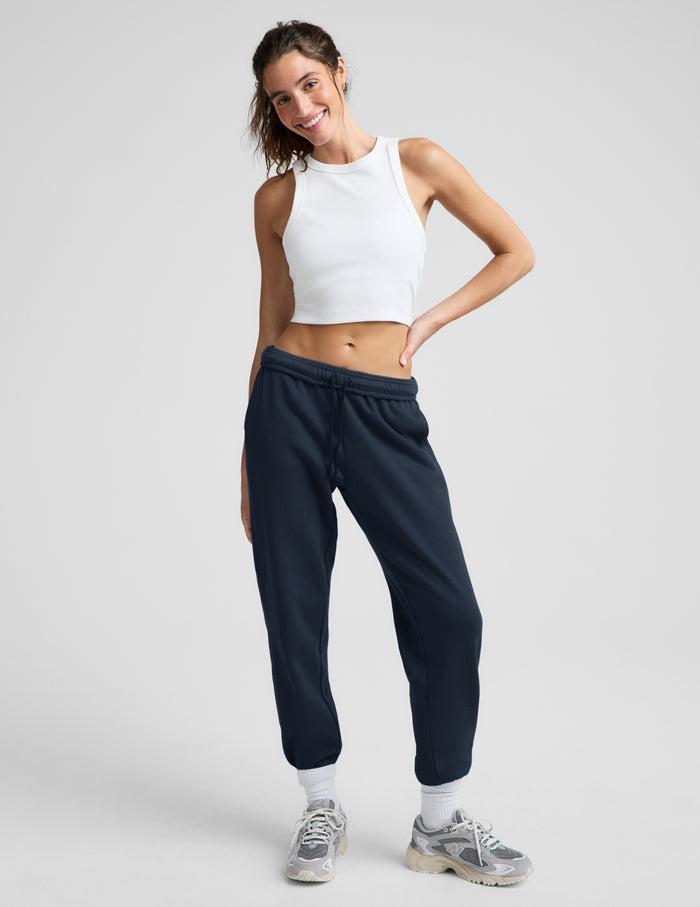 Woman wearing a white ribbed tank top and blue joggers against a plain background.