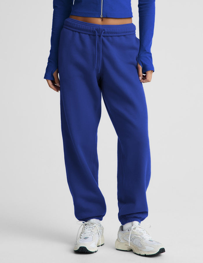 A person wearing bright royal blue sweatpants with a drawstring waist, paired with white sneakers, shown from the waist down against a light gray studio background.